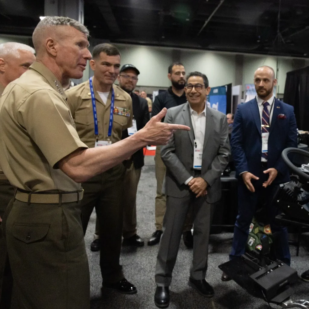 A Marine officer speaks to a group of men, both military and civilian, at an indoor event.